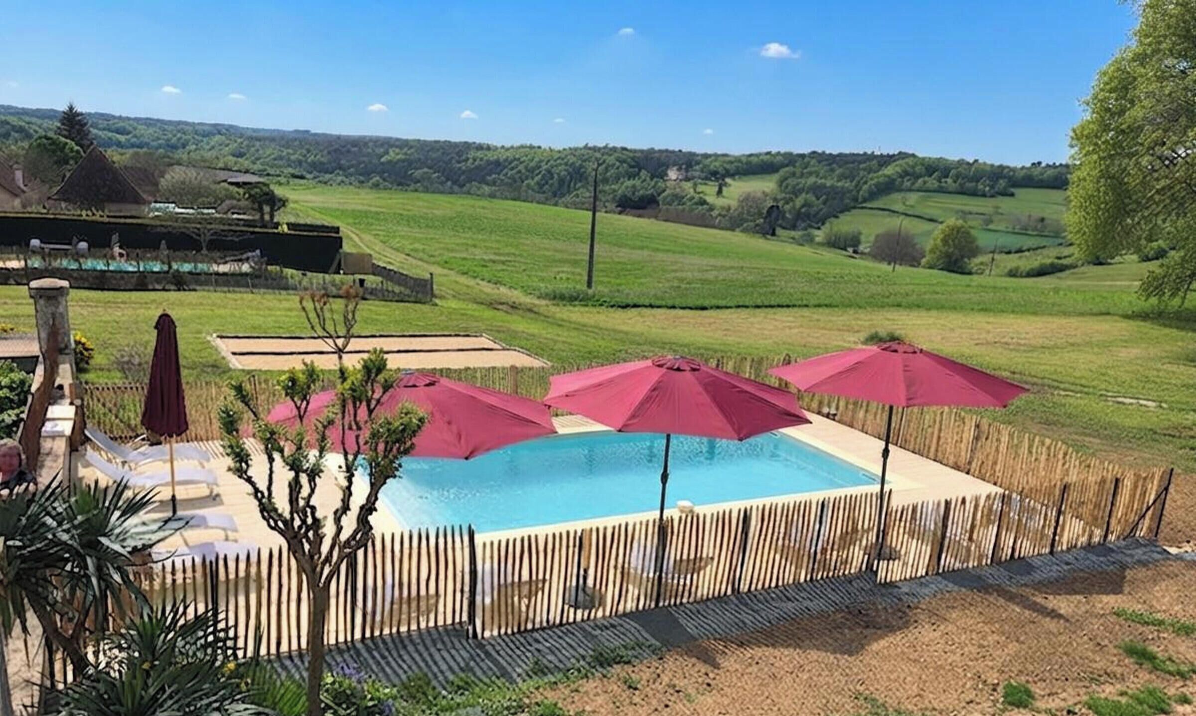 A swimming pool with a wooden fence, surrounded by umbrellas, set against a rural landscape.