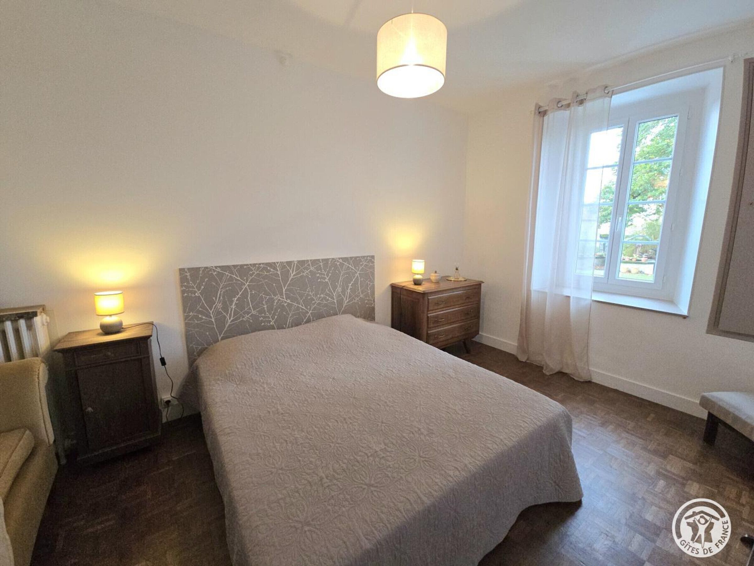 A bedroom with a bed, bedside lamps, a nightstand, a window with curtains, and a radiator.