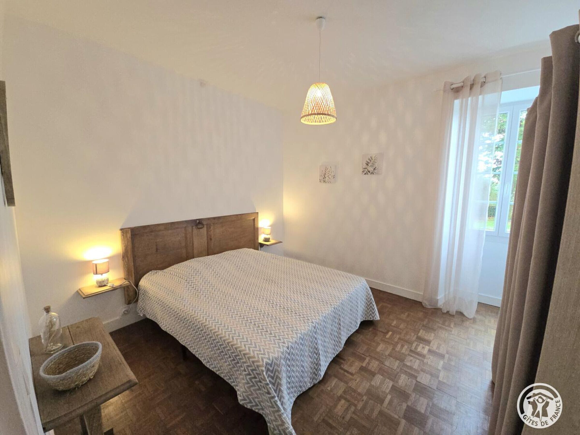 Une chambre à coucher avec une tête de lit en bois, un lit recouvert d’une couvre-lit à motifs, des lampes de chevet, une petite table avec un panier, et une fenêtre avec des rideaux.