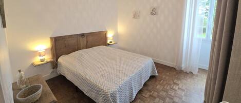 Une chambre à coucher avec une tête de lit en bois, un lit recouvert d’une couvre-lit à motifs, des lampes de chevet, une petite table avec un panier, et une fenêtre avec des rideaux.
