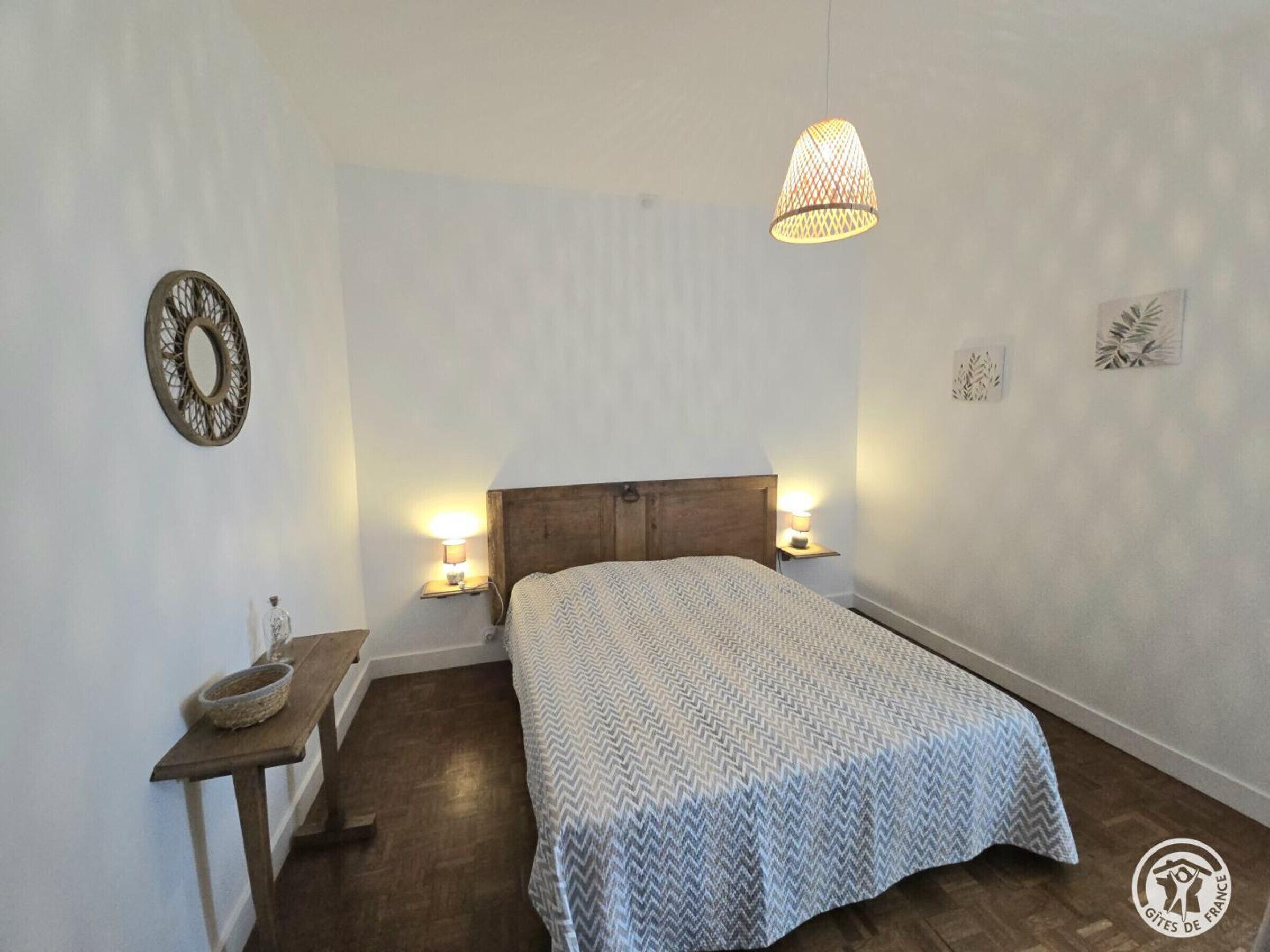 Ein Schlafzimmer mit einem Holzkopfteil, zwei Nachttischlampen, einem Bett mit gemustertem Bettbezug, einem kleinen Nachttisch mit einer Schale und einer Pendelleuchte.