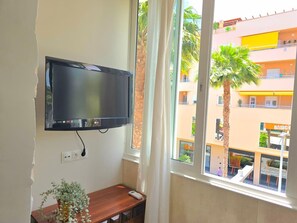 A room with a mounted TV, a small wooden table with a plant, and a window overlooking a building with balconies and palm trees. - Modern studio in central Torremolinos (Torremolinos)