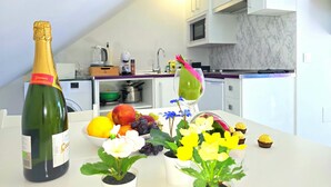Une cuisine avec des meubles blancs, un four à micro-ondes et un évier. On peut voir une bouteille de vin, un saladier de fruits et des plantes en pot sur le comptoir.