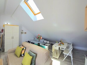 A modern living room with a high ceiling and skylight, a sofa with cushions, a dining table with chairs, and a small kitchen area.