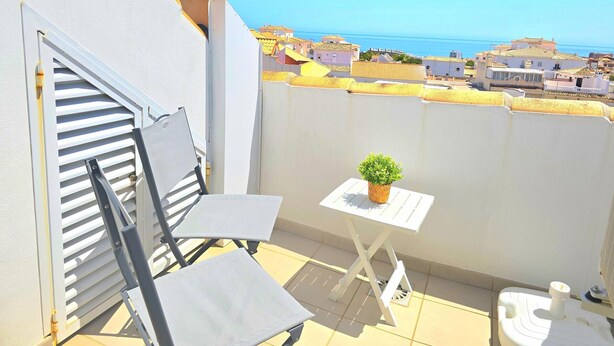 Un balcon avec une chaise, une petite table avec une plante et des toilettes. La vue comprend une ville côtière et des bâtiments aux toits jaunes.