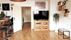 Een moderne woonkamer met houten vloer, een televisie op een houten dressoir, een eethoek met barkrukken en een gang die naar een andere kamer leidt.