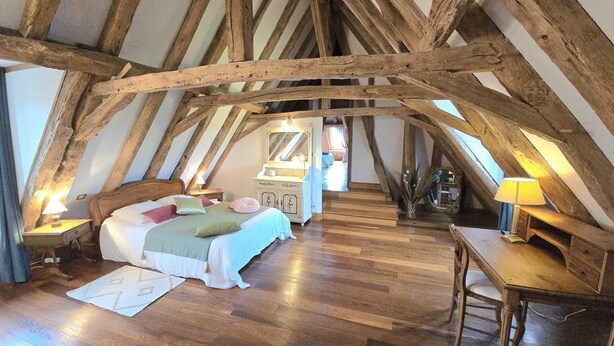 Ein Schlafzimmer mit schrägen Wänden, Holzbalken, einem Bett mit Kissen, einer Kommode und einem Schreibtisch mit Lampe.