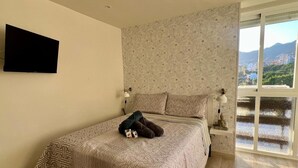 Un dormitorio con cama, televisor montado en la pared, ventana con vista a la ciudad y mesita de noche con lámpara.