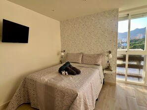 A bedroom with a bed, a TV mounted on the wall, a window with a view of a cityscape, and a nightstand with a lamp. - Modern Studio Benalmádena with Views (Marbella)