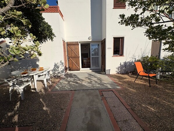 A building with a white exterior, a wooden door, and a small patio area with a table and chairs. - Ground Floor T3 Quiet 200m Beach, Pets Ok, Parking (Le Barcarès)