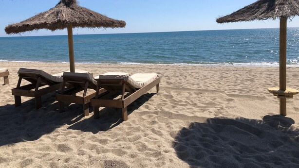 En strand med träsolstolar och parasoller med halmtak.