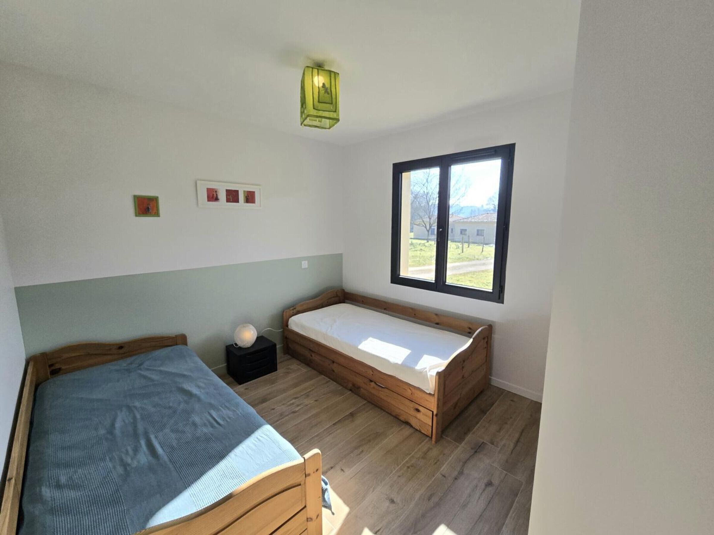 A small, bright room with two wooden beds, a window with a view of the outside, and a wall-mounted light fixture.
