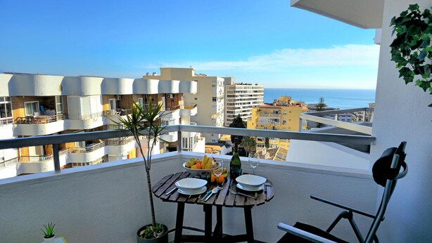 Un balcon avec une table dressée pour deux personnes, une plante en pot et une vue sur la mer et les bâtiments.