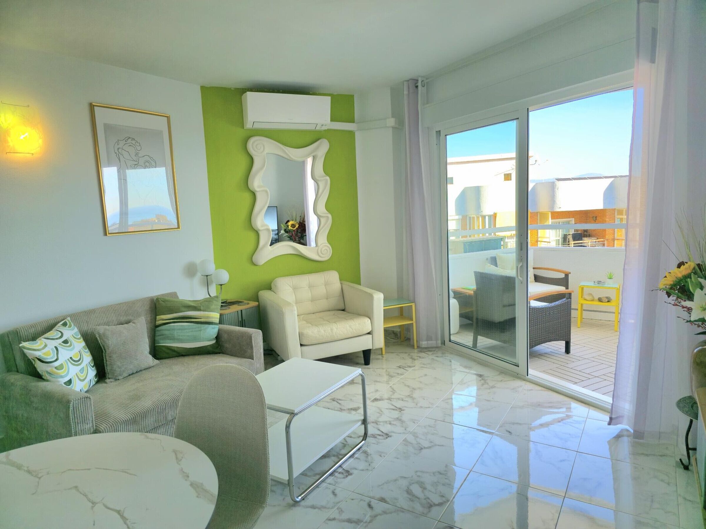 A modern living room with a green accent wall, a white sofa, a beige armchair, and a glass door leading to an outdoor balcony.
