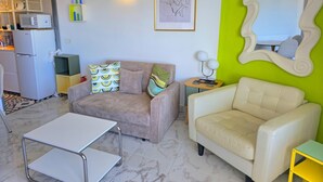 Un salon avec un canapé, un fauteuil blanc, une petite table et, au fond, un coin cuisine.