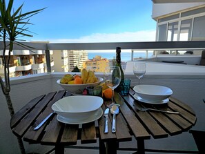Une table dressée avec des assiettes, des couverts, une bouteille et un saladier de fruits, placée sur un balcon donnant sur un paysage urbain.