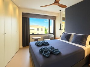 A modern hotel room with a large bed, a ceiling fan, a small round table, and a view of buildings through the window.
