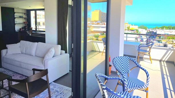 Een moderne woonkamer met een witte bank, een eettafel met stoelen en een balkon met rieten stoelen in blauw en wit.