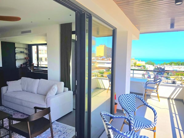 A modern living room with a white sofa, a dining table with chairs, and a balcony with blue and white wicker chairs.