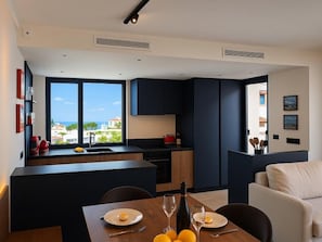 A modern kitchen with dark cabinetry, a dining area with a table set for two, and a view of the sea through large windows.