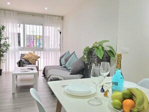 A modern living room with a dining table set for two, a sofa, and a window with sheer curtains. - Elegant 3-Bedroom Apartment in Torremolinos - Wifi and Air Conditioning (Torremolinos)