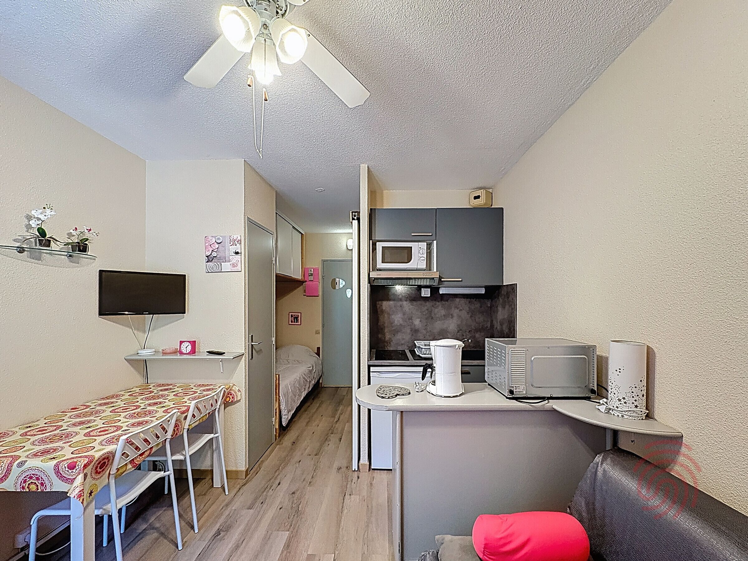 Ett kompakt bostadsutrymme med ett litet kök, en matplats med bord och stolar, en TV och en takfläkt.