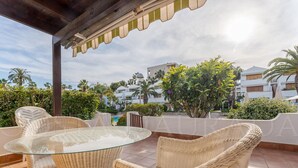 Une terrasse extérieure aménagée avec des meubles en osier, une table en verre et une vue sur une piscine et des bâtiments en arrière-plan.