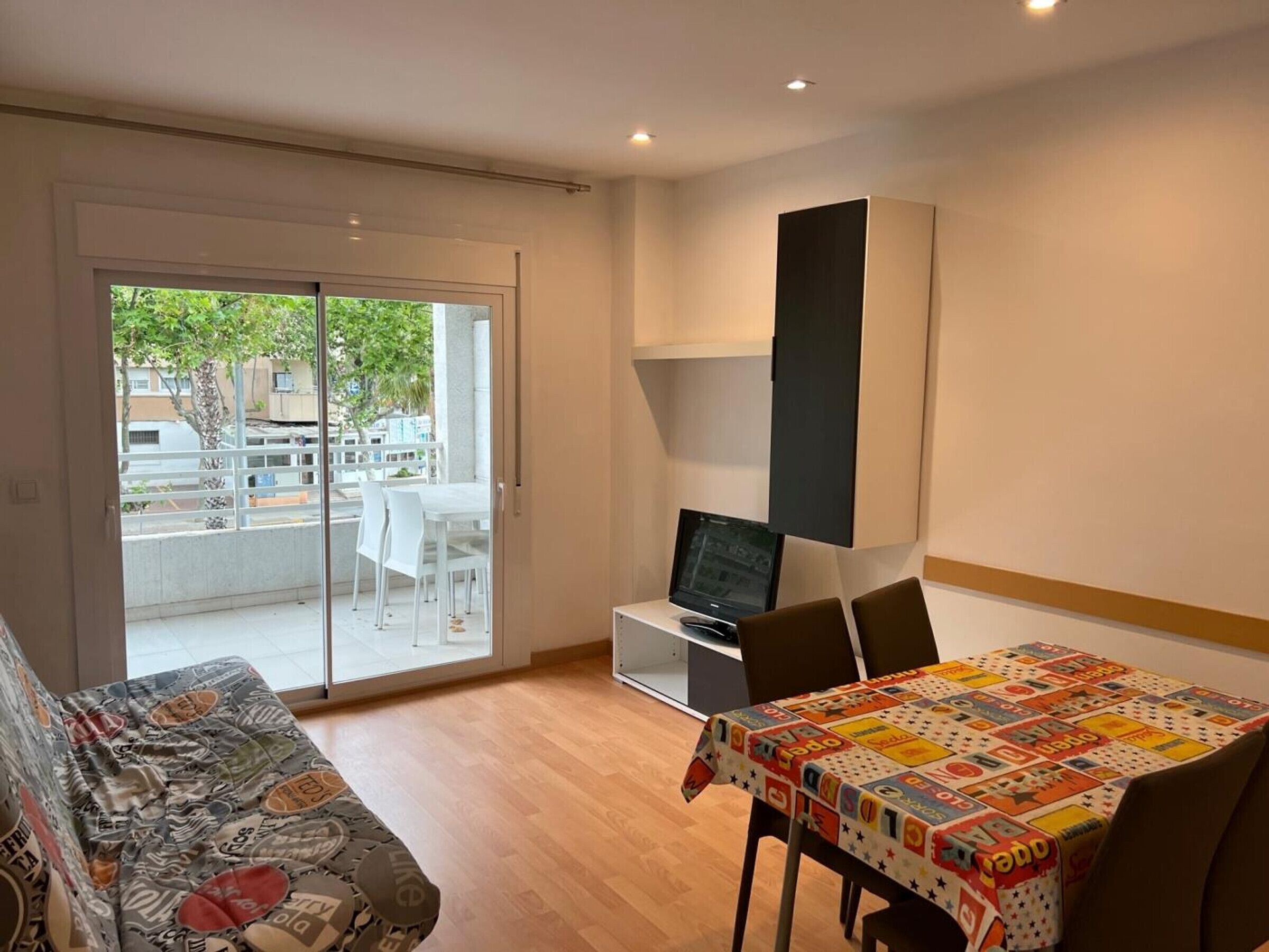 Un espace de vie compact doté d’une porte-fenêtre coulissante donnant sur un espace extérieur, d’une petite table recouverte d’une nappe colorée et d’un canapé agrémenté d’un revêtement à motifs.