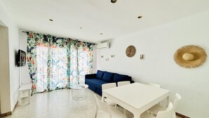 Een kamer met een witte tafel, witte stoelen, een blauwe bank, een gordijn met een patroon en een hoed die aan de muur hangt.