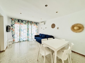 A room with a white table, white chairs, a blue sofa, a patterned curtain, and a wall-mounted hat. - Beachside Retreat: 2nd Line, A/C, WiFi, Balcony, Parking, Near Shops, Sleeps 6 (Roses)