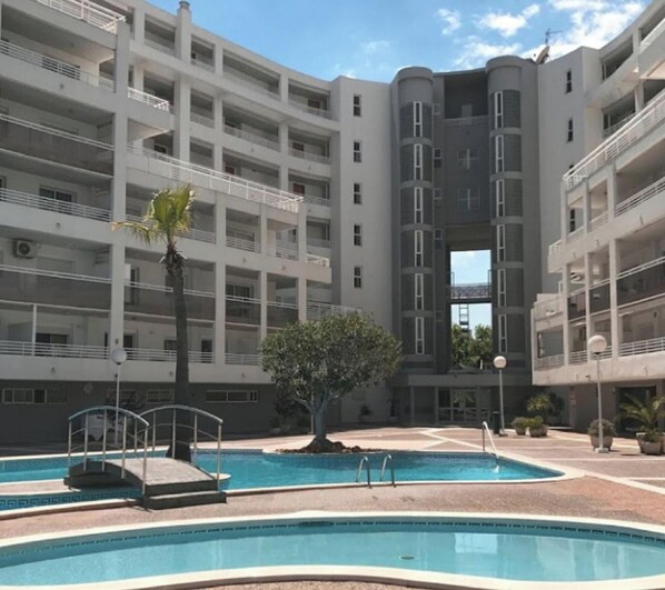 A modern multi-story building with a swimming pool, palm trees, and a clear sky. - Royal Apartments: Modern Stay with Pool, Terrace & A/C, 500m from Levante Beach, Near Port Aventura (Salou)