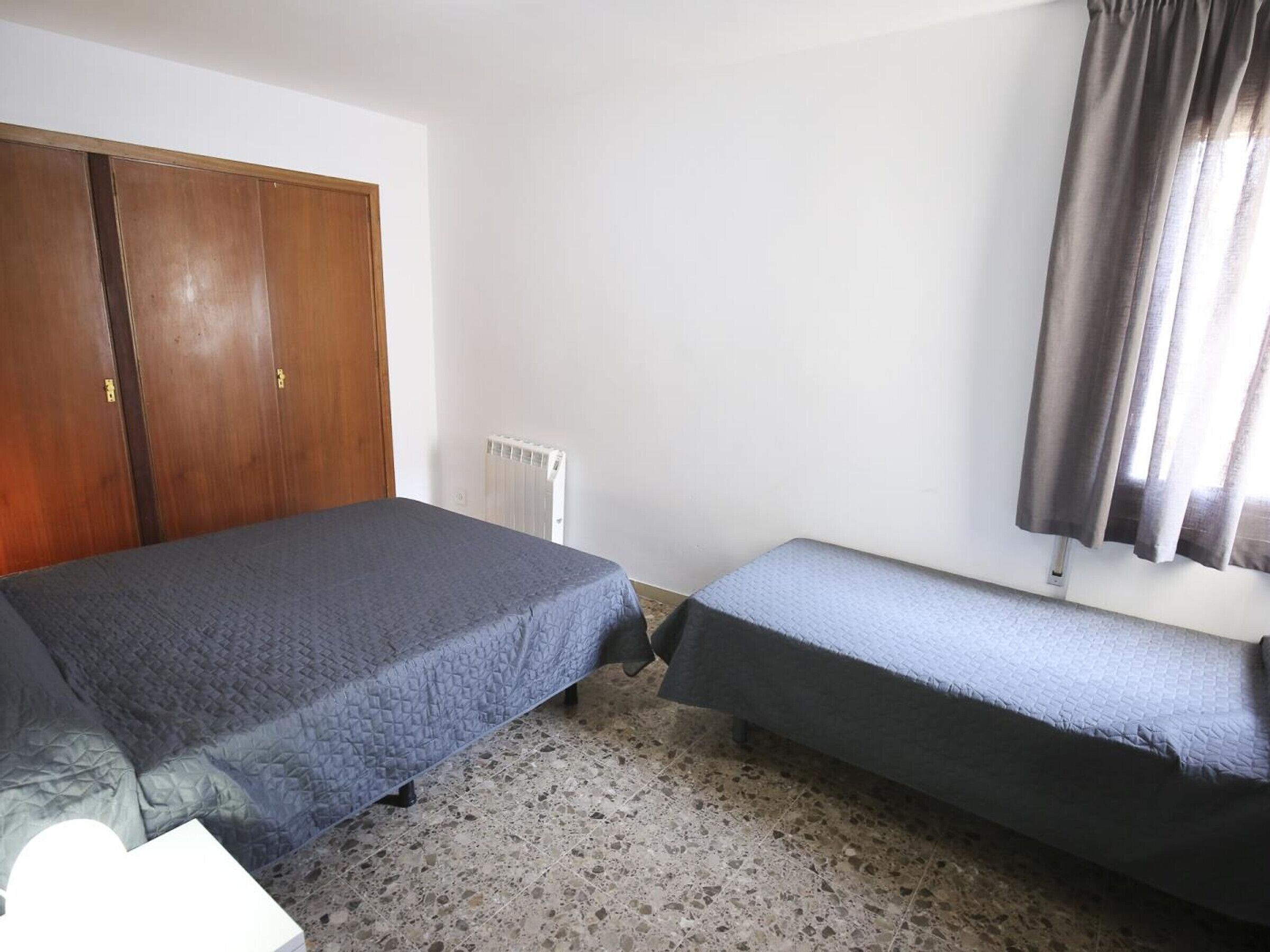 A bedroom with two beds, a wardrobe, a radiator, and a window with curtains.