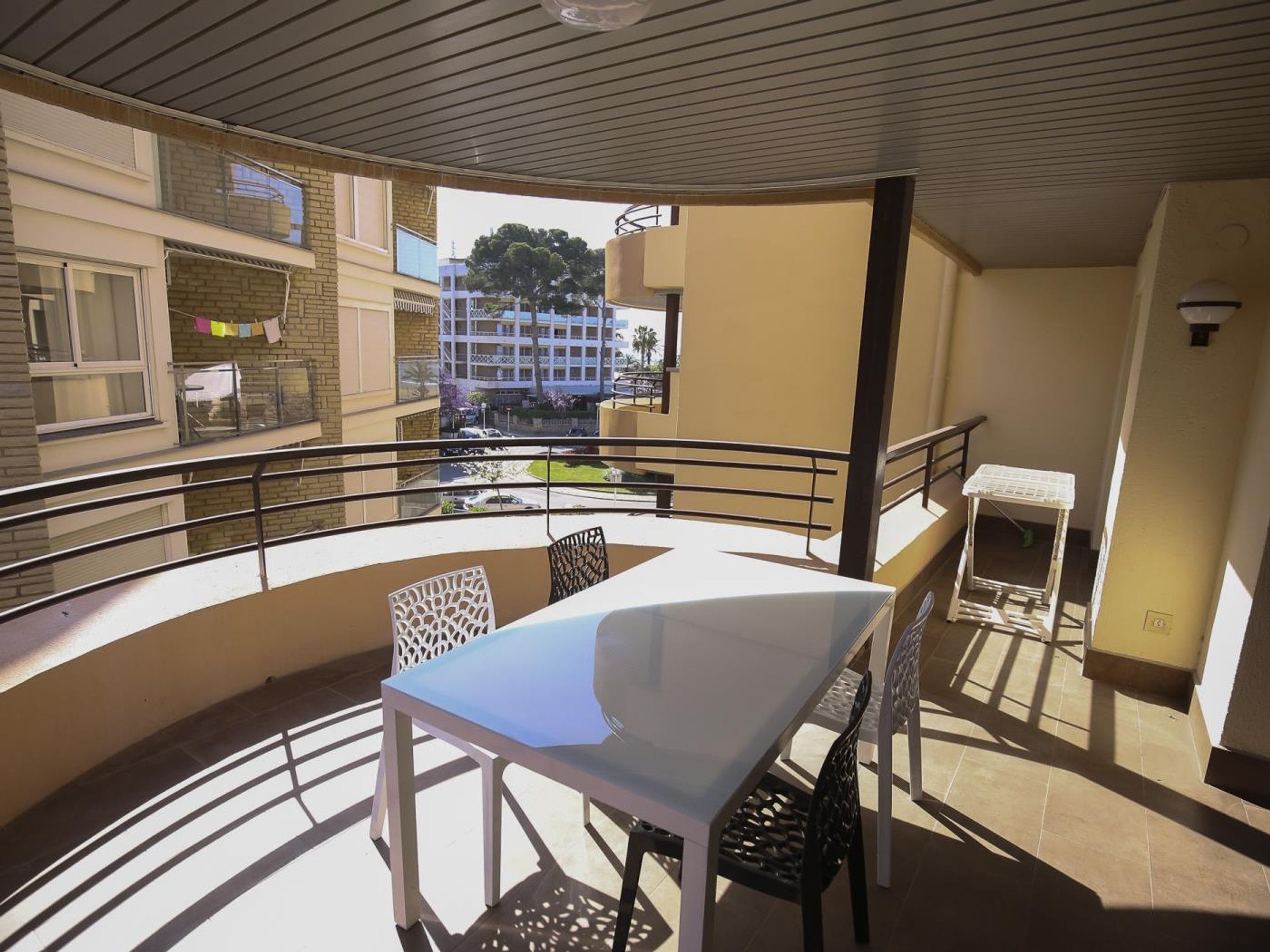 Un balcon avec une table et des chaises blanches, une petite table avec une chaise, et une vue sur un immeuble et des arbres.