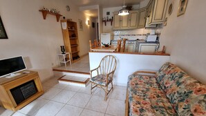 Una sala de estar con televisor, un sofá floral, una silla de madera y una escalera que conduce a una zona de cocina.