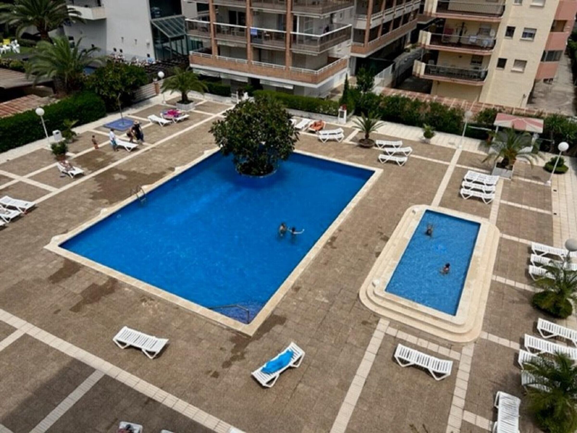 A rooftop pool area with two swimming pools, surrounded by lounge chairs and palm trees.