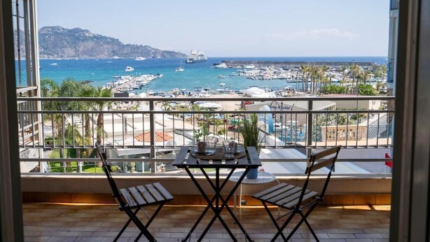 A balcony with a table and two chairs, overlooking a marina with boats and a cruise ship. - Naxos Seaside Escape: Terrace (Giardini Naxos)