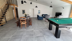 A spacious living area with a pool table, a dining table set for four, a staircase, and a TV in the background.