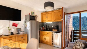 Una cucina compatta con mobili in legno, un forno a microonde integrato e una lavastoviglie. C'è una zona pranzo con tavolo e sedie, e un divano con cuscini decorativi. Un'ampia finestra offre vista su una zona boschiva.