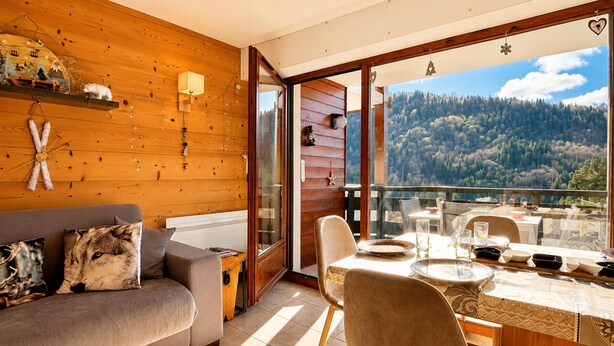 Arredamento in legno con divano, tavolo da pranzo e sedie, una porta vetrata che dà su un balcone con vista sulla foresta.