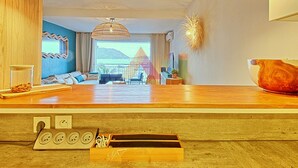 Un salon moderne avec un plancher en bois, une grande fenêtre et une vue sur la mer. On y trouve trois lampes suspendues, un canapé et une table basse.