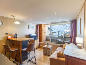 A cozy hotel room with a wooden desk, bar stools, a sofa, and a balcony with a view of snow-covered houses. - Cozy apartment 4 pers mountain decor near slopes (La Mongie)