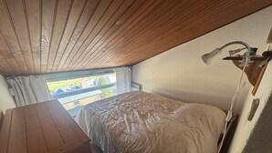 Ein kleiner, gemütlicher Raum mit Holzbalkendecke, einem Einzelbett mit weißer Bettdecke, einem kleinen Schreibtisch und einem Fenster mit Blick auf eine Wohngegend.