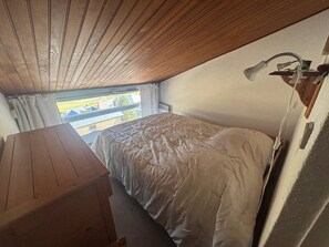 A small, cozy room with a wooden ceiling, a single bed with a white comforter, a small desk, and a window overlooking a residential area. - 3-Room Apartment in Les Angles with Balcony and Lake View (Les Angles)