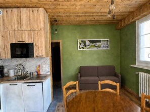 Eine kleine Küche mit Holzschränken, einer Mikrowelle und einem Spülbecken. Im Wohnbereich befindet sich ein braunes Sofa und ein Holzstuhl, und ein Gemälde ist an der Wand zu sehen.