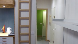 Kapea käytävä, jossa on puinen tikapuu, pieni keittiötila valkoisella työtasolla ja vihreä ovi käytävän päässä.