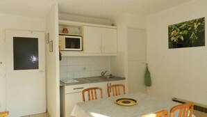 Une cuisine compacte avec des meubles blancs, un four à micro-ondes et un évier. On y trouve une table à manger recouverte d’une nappe à motifs et entourée de chaises en bois.