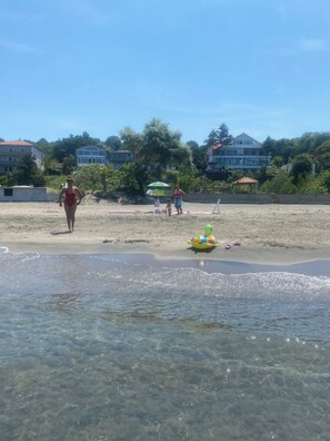 Egy tiszta vizű strand, egy úszóteknő-formájú úszóeszköz a vízben, és a háttérben épületek.