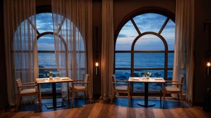 En matplats med två bord och stolar, stora fönster med utsikt över havet och ett trägolv.