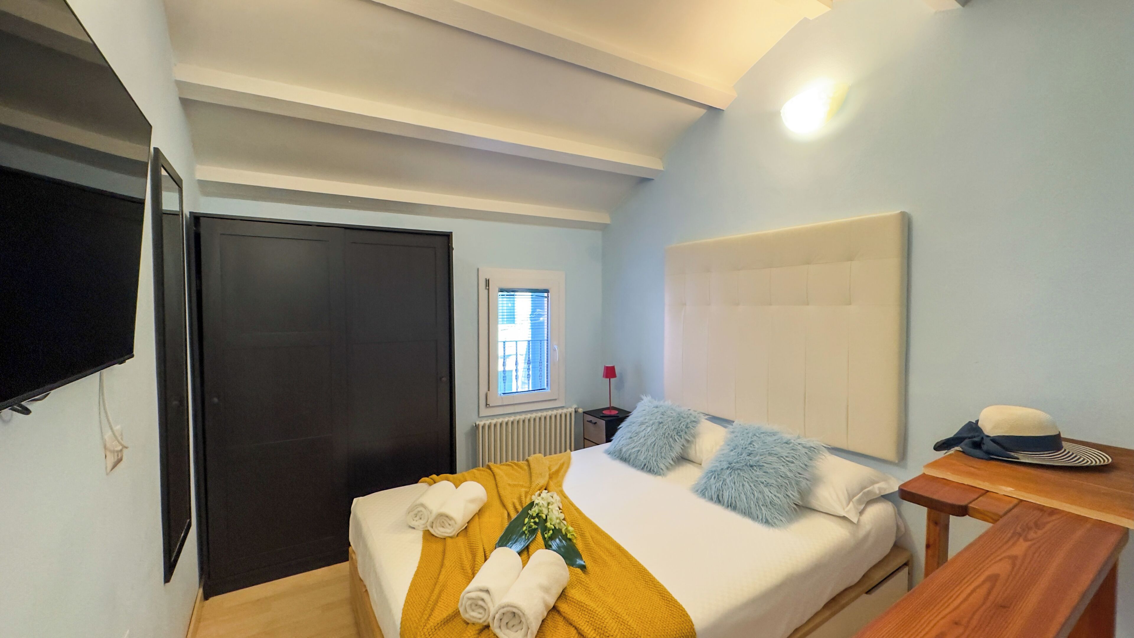 Un dormitorio con cama, televisor montado en la pared, ventana con persianas, mesita de noche y velador.
