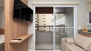 Un appartement moderne avec un téléviseur fixé au mur, un balcon offrant une vue sur les immeubles, un canapé beige et une petite étagère en bois sur laquelle repose une télécommande.